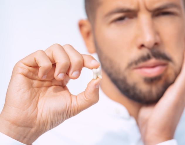 Wincing man holding his cheek with one hand and an extracted tooth in the other